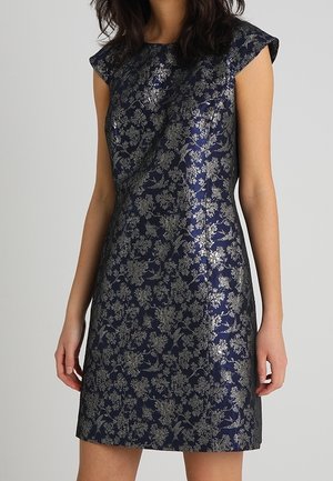 Mouwloze marineblauwe jurk met zilveren bloemenpatroon, getailleerde bolero, staande kraag, gedragen door een persoon tegen een effen achtergrond.