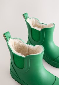 Gli stivali di gomma verdi presentano una morbida fodera in pelliccia beige e pannelli laterali in tessuto a trama. Hanno linguette al tallone per facilitarne la calzata.