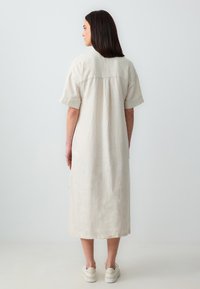 Beige linnen jurk met korte mouwen en een ontspannen pasvorm, met een achterste rugnaad en zijsplitten. Gepaarde met witte sneakers.