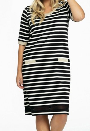 Kobieta w czarno-białej sukience w poziome paski do kolan, z złotym wykończeniem na rękawach, dekolcie i detalach kieszeni.