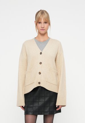 Beige ullkofta med V-hals, fem bruna knappar, oversized passform och framfickor. Bärs över en grå T-shirt och tillsammans med en svart kjol.