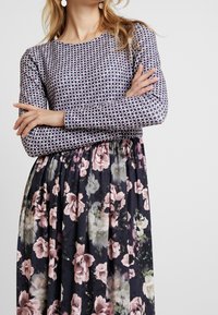 Vrouw in een gestreepte lange mouwtop en bloemenrok met gekruiste armen, tegen een effen lichte achtergrond.