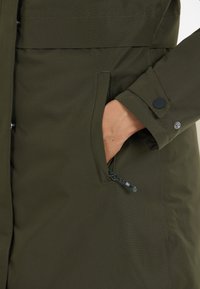 Olivgrön parkas med en slät textur, har en sidoficka och knappar på ärmarna. Designen är funktionell och strömlinjeformad.