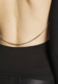Top in tessuto nero con schiena scoperta, caratterizzato da una catena argentata con pietre trasparenti lungo la parte bassa della schiena, che evidenzia il design minimalista e la texture.