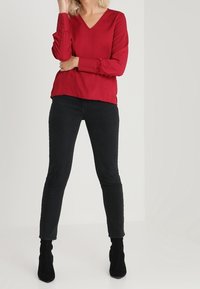 Rød v-hals bluse med lange ærmer, der har knapdetaljer ved manchetterne, parret med slanke sorte bukser og sorte ankelstøvler.