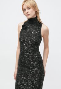 Knit sleeveless dress in dark gray, featuring a high turtleneck and scattered metallic embellishments. Accented with a floral brooch.