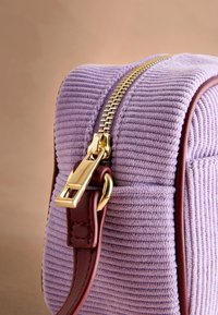Fialová kordurová taška se zlatým zipem a vínovým koženým popruhem. Má vertikální žebrování a kompaktní, obdélníkový tvar.