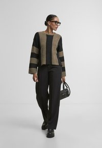 Czarny i brązowy w paski krótki sweter z teksturowanym splotem, w zestawieniu z czarnymi spodniami o wysokim stanie i masywnymi czarnymi butami, trzymając małą czarną torbę.