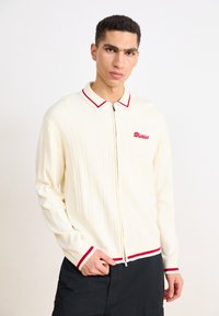 Krēmkrāsas ribota adīta zip-up džemperis ar sarkanu maliņu apkaklei un piedurknēm, ar vārdu "Dickies" izšūtu sarkanā krāsā kreisajā krūtī.