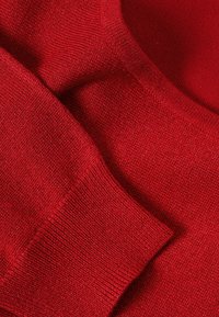 Pull en tricot rouge avec une texture douce, un col arrondi et des poignets côtelés. Le tissu présente une légère variation de densité de tricot et de brillance.