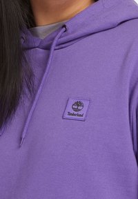 Primo piano di una persona che indossa una felpa con cappuccio Timberland viola con coulisse e una maglietta grigia sotto.