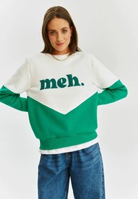 Zelená a bílá mikina s texturou s nápisem "meh.", chevronovým vzorem a jemnou tkaninou. V kombinaci s modrými džínami se zvýšeným pasem.