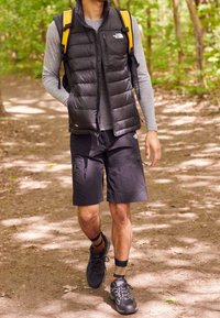 Černá péřová vesta přes šedou dlouhýrukávovou košili, doplněná černými kraťasy. Černé turistické boty s vzorovanými ponožkami. Zelená lesní cesta v pozadí.