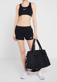 Sujetador deportivo y pantalones cortos negros de Nike, combinados con una bolsa de deporte negra grande con dos asas y un bolsillo frontal. Logo de Nike en blanco visible.