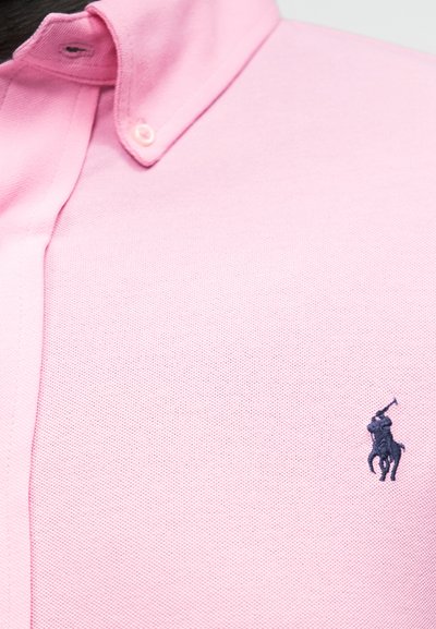 Nærbilde av en rosa poloshirt med knappet krage og en liten mørk brodert polospillerlogo på brystet.