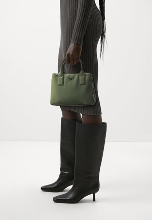 Personne portant une robe grise côtelée et des bottes noires à talons montant jusqu'aux genoux, tenant un petit sac à main vert avec deux anses.