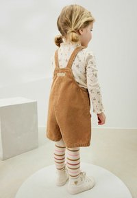 Salopette di velluto a coste marrone e una camicia a maniche lunghe con fantasia floreale abbinate a calze a righe fino al ginocchio e sneakers bianche.