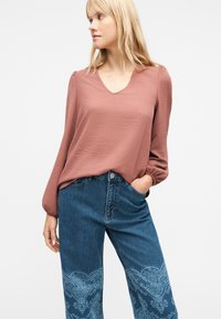 Ξανθιά γυναίκα που φοράει μια υφασμάτινη, μακρυμάνικη μπλούζα σε απόχρωση ξεθωριασμένου ροζ και ψηλόμεσα μπλε τζιν με ανοιχτόχρωμα μπλε λουλουδάτα σχέδια.