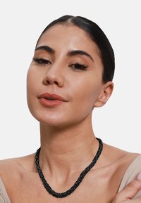 Zwarte gevlochten ketting met een gestructureerd ontwerp, gedragen op een blote schouder. Het materiaal lijkt geweven of koordachtig, met een flexibele vorm.