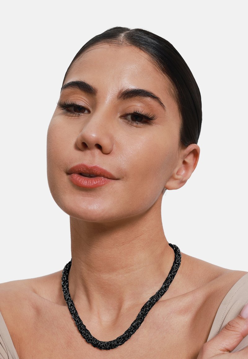 Zwarte gevlochten ketting met een gestructureerd ontwerp, gedragen op een blote schouder. Het materiaal lijkt geweven of koordachtig, met een flexibele vorm.
