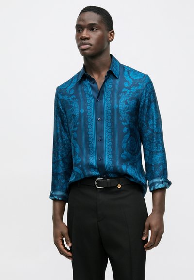 Kék mintás ing bonyolult virágmintákkal, hosszú ujjú és elején gombokkal, fekete nadrággal és övvel párosítva.