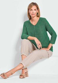 Groene gebreide pullover met een v-hals en een textuurpatroon, gecombineerd met lichtbeige tapered broek en tan kleurige sandalen met hakken en knoopdetails.