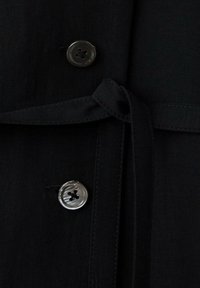 Deux boutons noirs et une ceinture en tissu nouée en un nœud sur un vêtement noir, montrant les détails des coutures.
