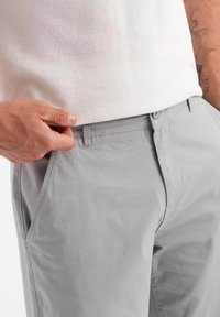Gros plan d'une personne portant un pantalon gris clair et une chemise blanche texturée, une main ajustant la taille du pantalon.