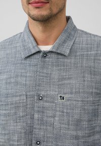 Grijze, gestructureerde shirt met een kraaghals. Voorzien van donkere knopen, twee voorzakken en een klein logopatch.