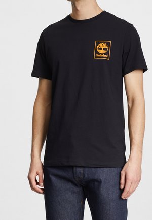 Mann trägt ein schwarzes Timberland-T-Shirt mit einem orangefarbenen Baumlogo auf der linken Brustseite und dunkelblaue Jeans, steht vor einem schlichten Hintergrund.
