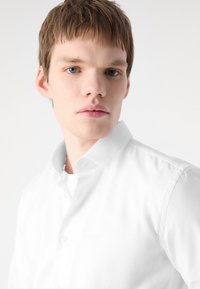 Weißes Hemd mit Knöpfen und strukturiertem Muster sowie spitzem Kragen, getragen über einem weißen Unterhemd. Das Gesicht des Modells ist sichtbar und zeigt Neutalität.