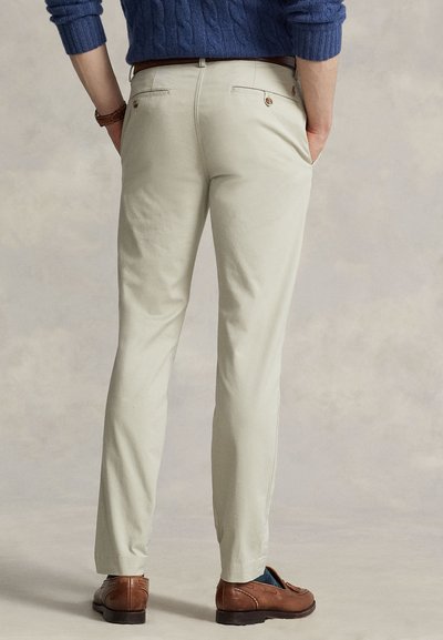 Moški, ki nosi bež hlače po meri, rjave usnjene loaferje in modro pulover z vzorcem kitke, stoji z rokami v žepih pred nevtralnim ozadjem.