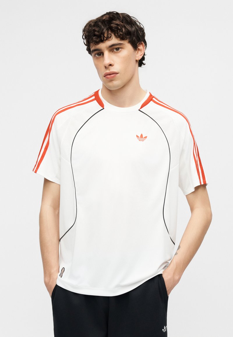 Junger Mann mit lockigem Haar, der ein weißes Adidas-Shirt mit orangefarbenen Streifen an den Ärmeln und schwarze Hosen trägt, steht mit den Händen in den Taschen.