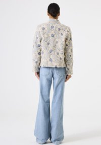 Frau mit dunkelhaariger Hochsteckfrisur, die eine strukturierte blumenbedruckte Jacke in Creme und Blau sowie weite, hellblaue Jeans trägt, steht vor einem weißen Hintergrund.