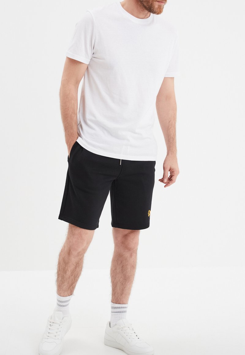 Pantalones cortos de algodón negros con una cinturilla elástica, que presentan un pequeño logo amarillo. Combinados con una camiseta blanca de manga corta y zapatillas blancas.