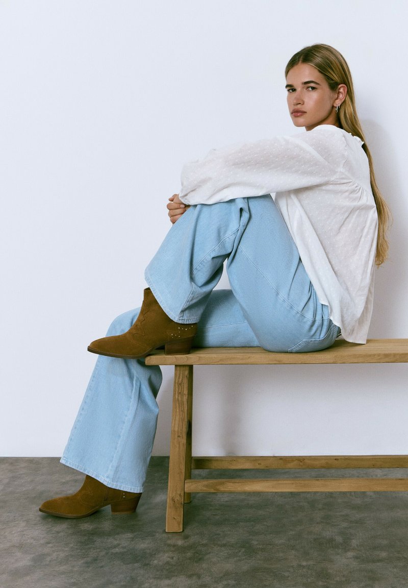 Jeans de pierna ancha azul claro, blusa blanca texturizada y botas de ante marrón hasta el tobillo con acentos decorativos, sentada en un banco de madera frente a un fondo sencillo.