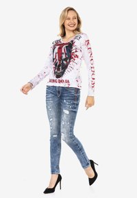 Weißes Langarmhemd mit grafischem Totenkopfdesign, akzentuiert mit roten Spritzern, kombiniert mit ausgebleichten blauen Jeans und schwarzen High Heels.