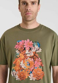 Camiseta de algodón verde oliva con estampado floral en rosa, naranja y azul con detalles de contorno en negro, corte relajado, cuello redondo.