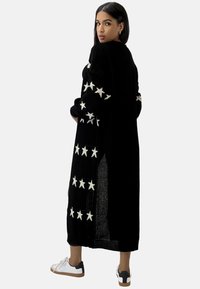 Cardigan long en tricot noir avec des motifs d'étoiles blanches sur les manches, à coupe ample et avec des fentes sur les côtés pour plus de confort.