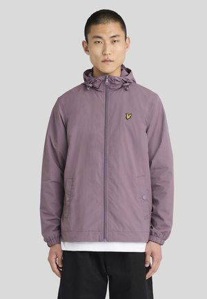 Homme portant une veste à capuche mauve zippée avec des poignets élastiques et des poches avant, par-dessus une chemise blanche et un pantalon noir.