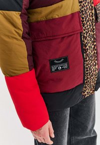 Osoba oblečená v mnohofarebnej prešívanej bunde s červenými, marhuľovými, olivovými a leopardími vzormi, pričom na vrecku sa nachádza čierny nášivka s textom.