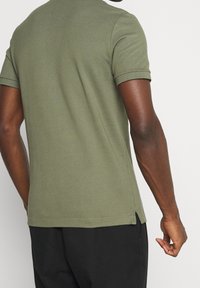 Polo en coton vert olive avec un design classique, manches courtes, fentes latérales, col et poignets côtelés. Texture lisse, coupe décontractée.