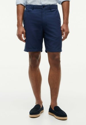 Mann trägt dunkelblaue, maßgeschneiderte Shorts, hellblaues Hemd und dunkelblaue Slipper mit geflochtenen Sohlen, steht auf weißem Hintergrund.