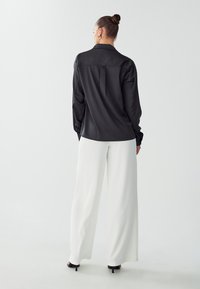 Blusa nera in satin con maniche lunghe e colletto, abbinata a pantaloni bianchi a vita alta e gamba larga e sandali neri con tacco.