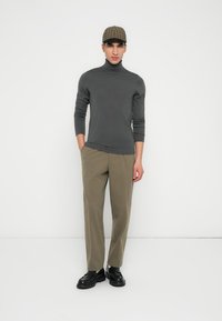 Mand iført en mørkegrå turtleneck, olivengrønne bukser, sorte sko og en brun ternet hat, der står foran en neutral hvid baggrund.