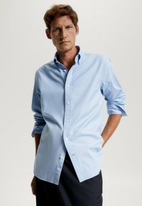 Camicia azzurra chiara con bottoni, realizzata in tessuto liscio, caratterizzata da un colletto classico, maniche lunghe arrotolate ai polsi e un orlo curvo.