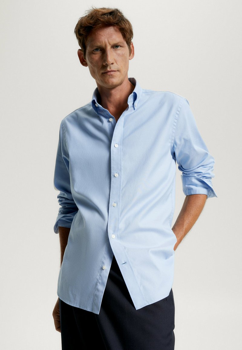 Camicia azzurra chiara con bottoni, realizzata in tessuto liscio, caratterizzata da un colletto classico, maniche lunghe arrotolate ai polsi e un orlo curvo.