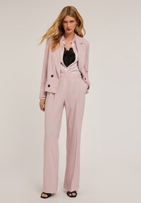 Roze op maat gemaakt pak met een dubbelzijdige blazer en hoge taille trouser. Zwarte cami-top onder een lichtroze shirt met een open kraag. Zwarte hakken.