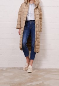 Béžový prošívaný dlouhý zateplený kabát s kapucí, dvěma předními kapsami a zipovým zapínáním. Nositý přes modré džíny a krémové platformové mokasíny.