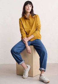 Musztardowy sweter z krótkim kołnierzykiem i guzikami; luźne niebieskie dżinsy; kremowe sneakersy z teksturowanym wzorem; drewniany postument.